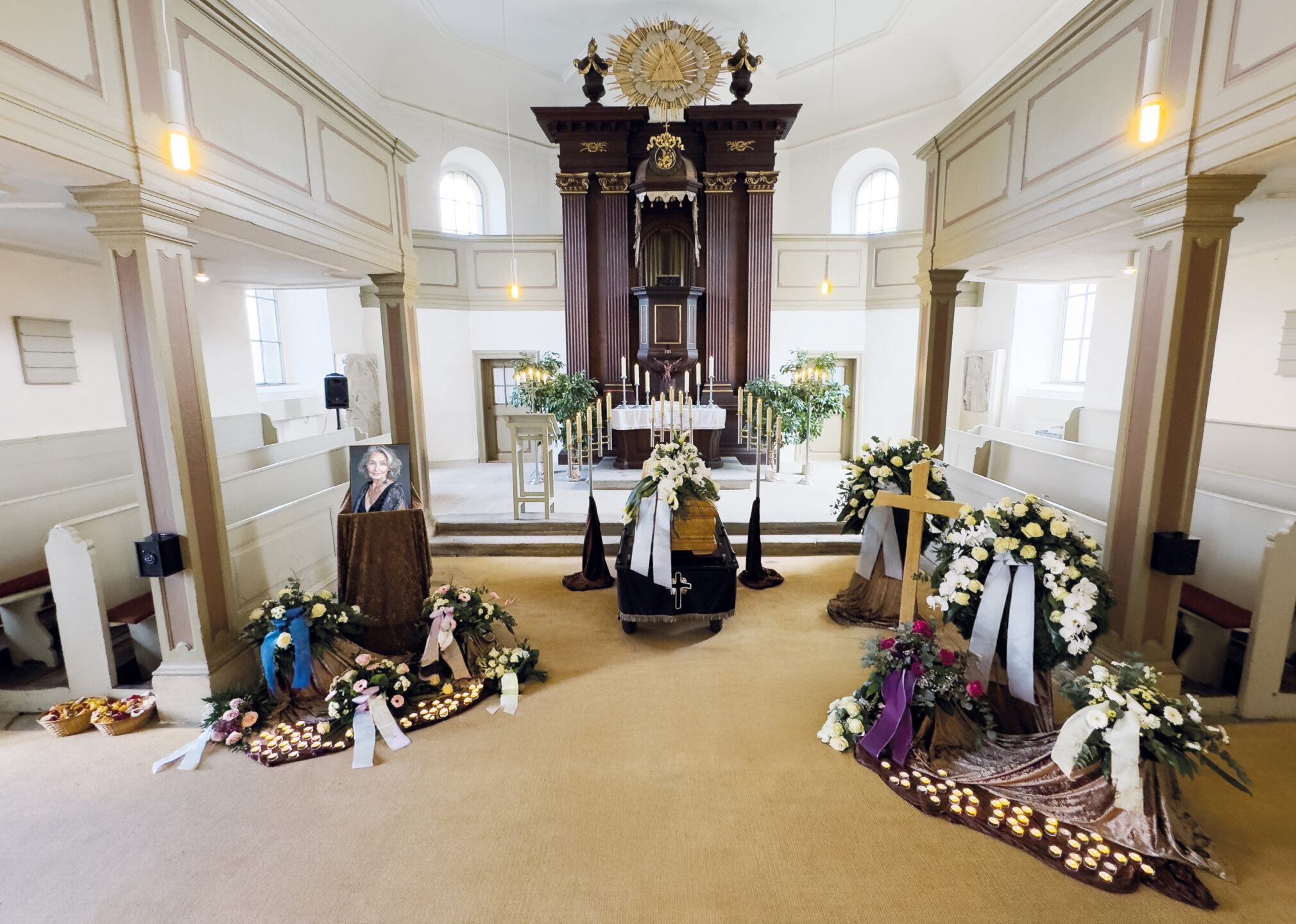 Ein für eine Beerdigung eingerichteter Kirchenraum mit einem Sarg in der Mitte, umgeben von Blumenschmuck, Kerzen, einem großen Kreuz und einem gerahmten Foto einer Frau. Licht fällt durch hohe Fenster ein.