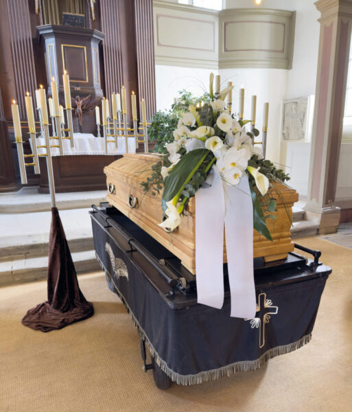 Ein hölzerner Sarg mit weißem Blumenschmuck und einer großen weißen Schleife steht auf einem Wagen, der mit einem schwarzen Tuch mit Kreuzmuster bedeckt ist, in einer mit Kerzen beleuchteten Kirche, die für die Trauerfeier vorbereitet ist.