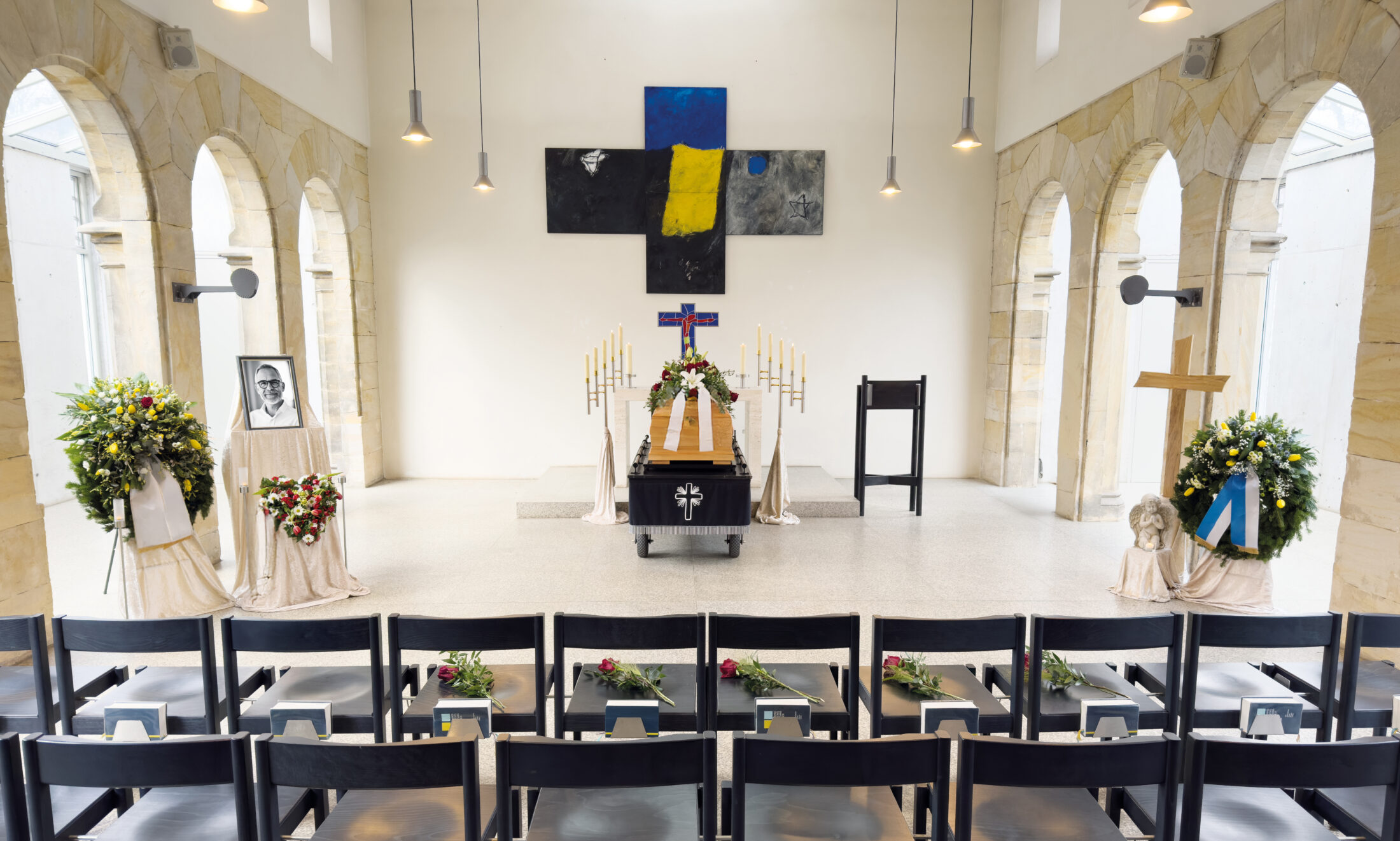Eine Beerdigungskapelle mit einem Sarg an der Stirnseite, umgeben von Kerzen, Blumenschmuck, gerahmten Fotos und einem Kreuz. Vor dem Altar, unter großen Bogenfenstern und Hängelampen, stehen Stuhlreihen mit einzelnen roten Rosen.
