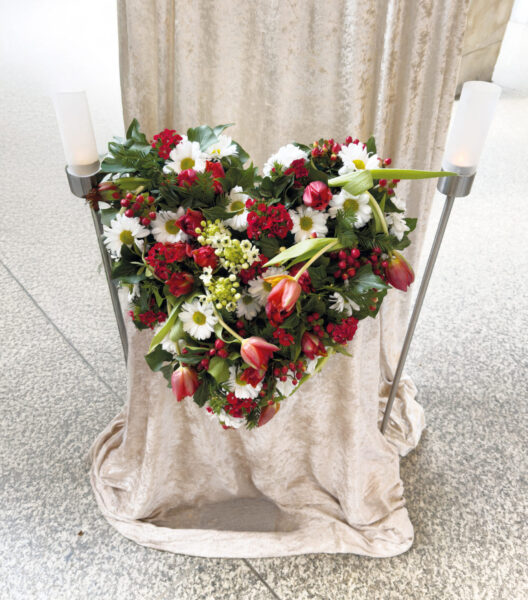 Ein herzförmiges Blumenarrangement mit roten, weißen und rosafarbenen Blüten hängt an einem beigen Stoffvorhang, flankiert von zwei hohen Kerzenhaltern mit weißen Kerzen - ein eleganter Tribut für eine Trauerfeier in der Nähe des Sargs auf einem grau gesprenkelten Boden.