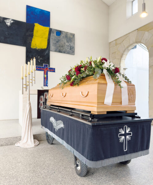 Ein hölzerner Sarg mit einem Blumenarrangement ruht auf einem fahrbaren Podest, das mit einem schwarzen Tuch mit weißem Kreuz bedeckt ist, in einer modernen Kapelle für eine Trauerfeier, mit einem abstrakten Kreuzkunstwerk und Kerzen im Hintergrund.