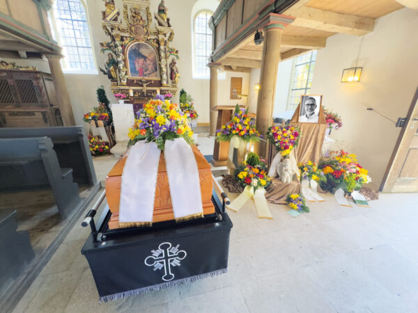 Ein mit weißen Bändern und bunten Blumen geschmückter Holzsarg liegt in einer Kirche, umgeben von Blumengestecken und Kränzen. Ein schwarzes Tuch mit einem weißen Kreuz bedeckt den Sargständer. Ein Porträt und weitere Blumen sind in der Nähe ausgestellt.