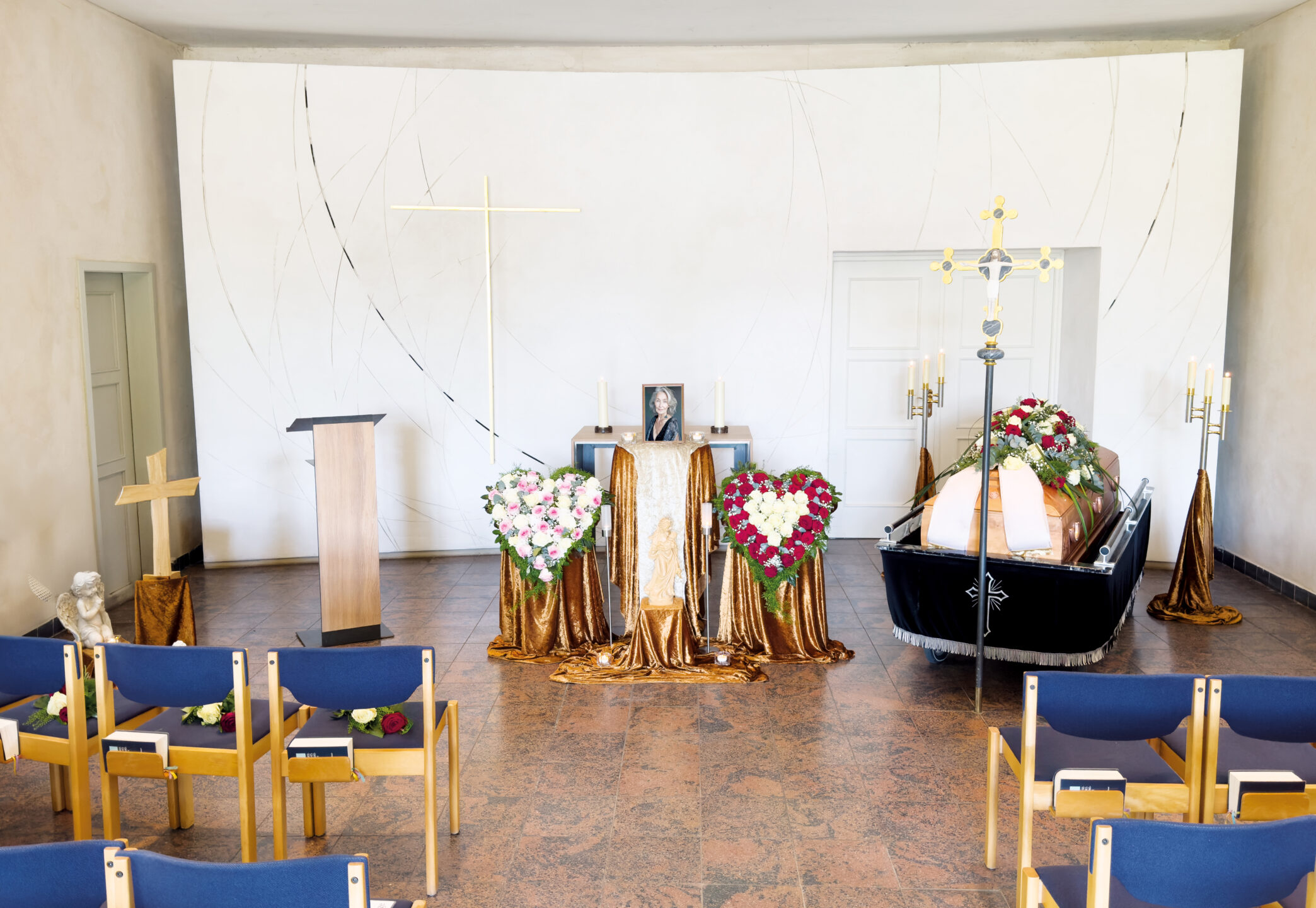 Eine Beerdigungskapelle mit blauen Stühlen vor einem Sarg, umgeben von Blumengestecken und goldenen Vorhängen. Ein Foto, ein Kreuz, Kerzen und ein Podium sind an der Stirnseite des Raumes angebracht.