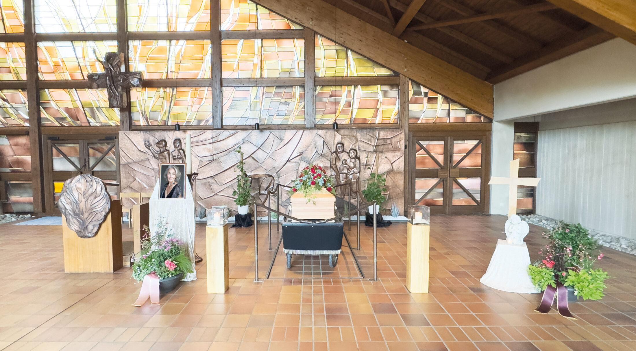 Ein Holzsarg mit Blumen darauf steht in der Mitte einer Kapelle, umgeben von Pflanzen, Kerzen, einem Kreuz, einem Foto und religiösem Dekor. Der Hintergrund besteht aus Buntglas und Holzvertäfelung.