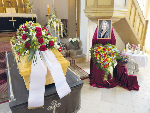 Ein hölzerner Sarg mit einem großen Blumenarrangement und weißen Bändern ist in einer Kirche aufgestellt. In der Nähe steht das Porträt einer lächelnden Frau neben weiteren Blumen und Kerzen, um eine feierliche Atmosphäre für die Trauerfeier zu schaffen.