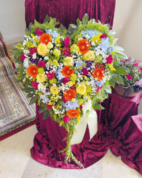Ein großes herzförmiges Blumenarrangement für eine Trauerfeier mit roten, orangefarbenen, gelben, violetten und blauen Blumen und Grünzeug ist auf tiefrotem Samt neben einem Sarg ausgestellt. Ein dekorativer Teppich und ein weiteres Blumenarrangement befinden sich in der Nähe.