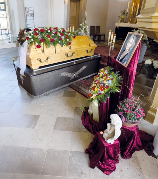 Eine Trauerfeier mit Sarg in einer Kirche, mit einem mit Blumen geschmückten Holzsarg, einem großen Blumenarrangement, einem gerahmten Porträt, Kerzen und einer kleinen Engelsstatue, während Licht durch die Fenster fällt.