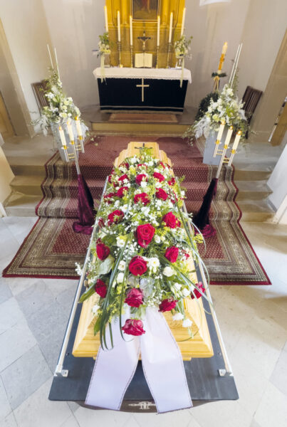Ein hölzerner Sarg, der mit einem großen Strauß roter und weißer Blumen geschmückt ist, steht an der Stirnseite der Kirche in der Nähe des Altars, umgeben von Kerzen und Blumenschmuck während der Trauerfeier.