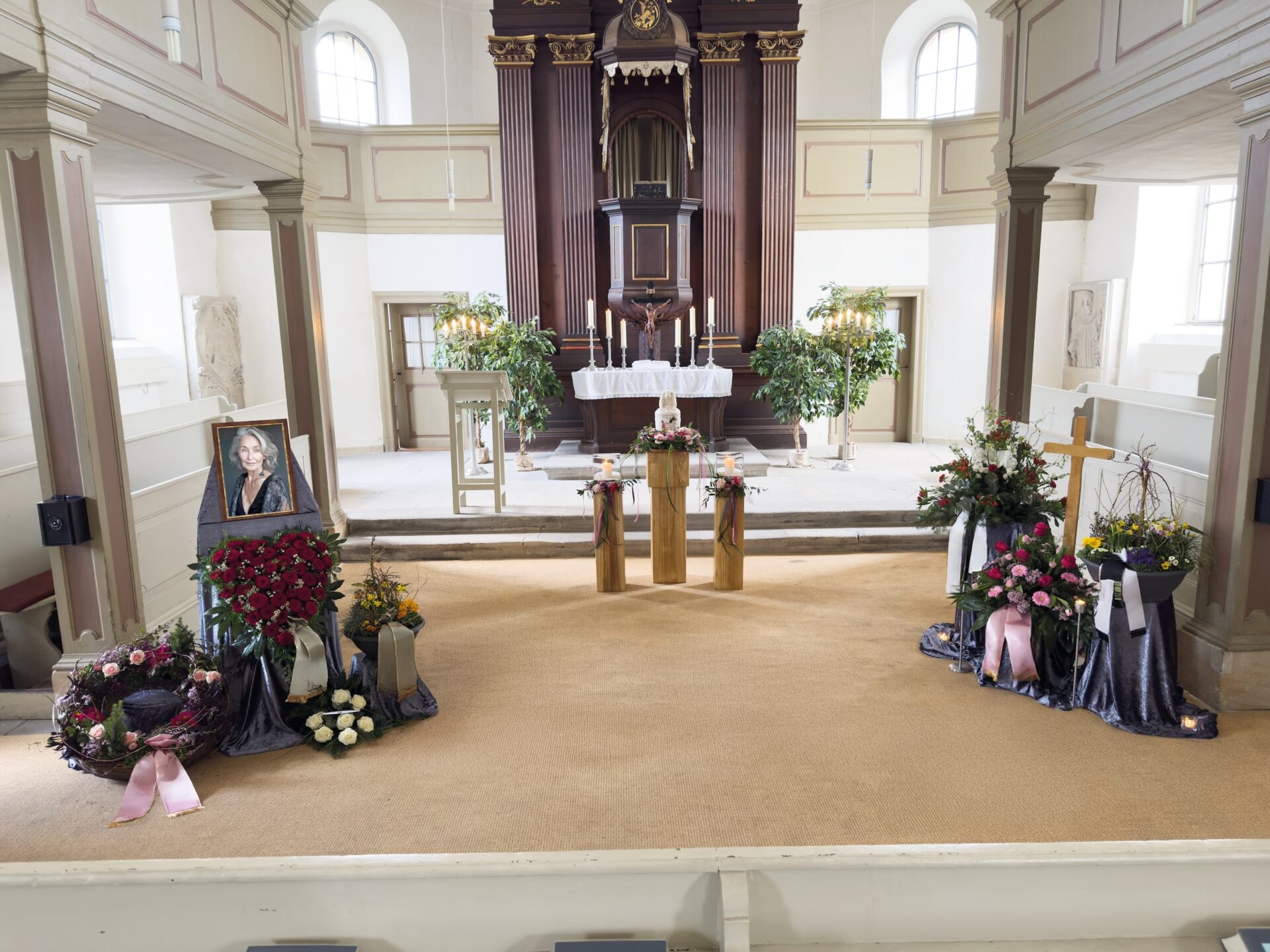 Ein für eine Urnentrauerfeier geschmückter Kirchenaltar mit Blumenarrangements, Kränzen, Kerzen, einem gerahmten Frauenporträt und einem Holzkreuz, durch dessen Fenster Tageslicht fällt.