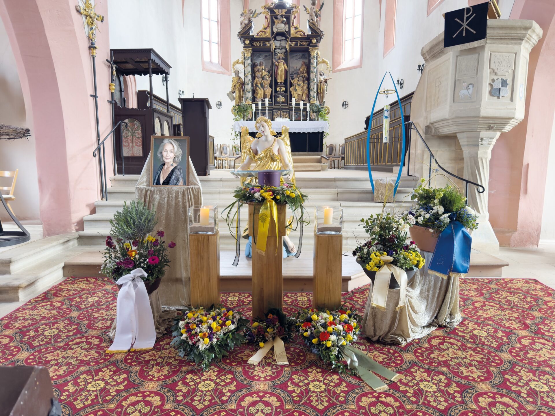 Ein Kirchenraum mit Blumenarrangements, Kerzen und einem gerahmten Porträt, das in der Nähe des verzierten Altars ausgestellt ist, lässt auf eine Urnentrauerfeier oder einen Trauergottesdienst schließen. Der Boden ist mit einem gemusterten roten Teppich ausgelegt, der die feierliche Atmosphäre noch verstärkt.
