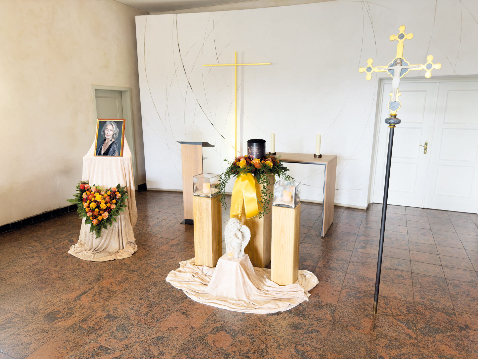 Bei einer Urnentrauerfeier steht eine schwarze Urne auf einem Sockel, umgeben von Blumen, Kerzen und einem gelben Tuch. Ein gerahmtes Foto des Verstorbenen und ein großes Kreuz sorgen für eine feierliche Atmosphäre in einem hellen Raum.