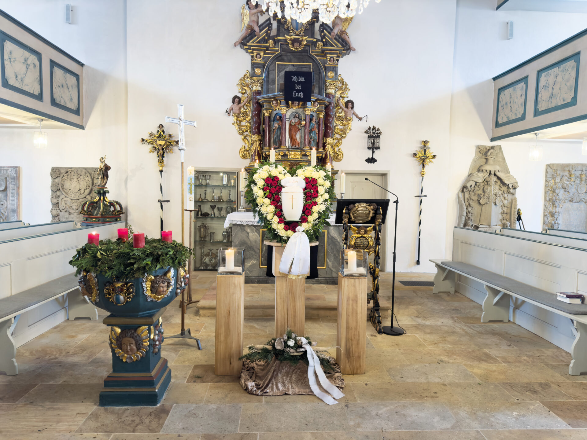 Ein Kirchenraum für eine Urnentrauerfeier mit einer geschmückten Urne auf einem Sockel, umgeben von Kerzen und einem großen Blumenkranz. Auf der linken Seite befindet sich ein Adventskranz mit roten Kerzen, während der Altar dahinter mit religiösem Schmuck verziert ist.