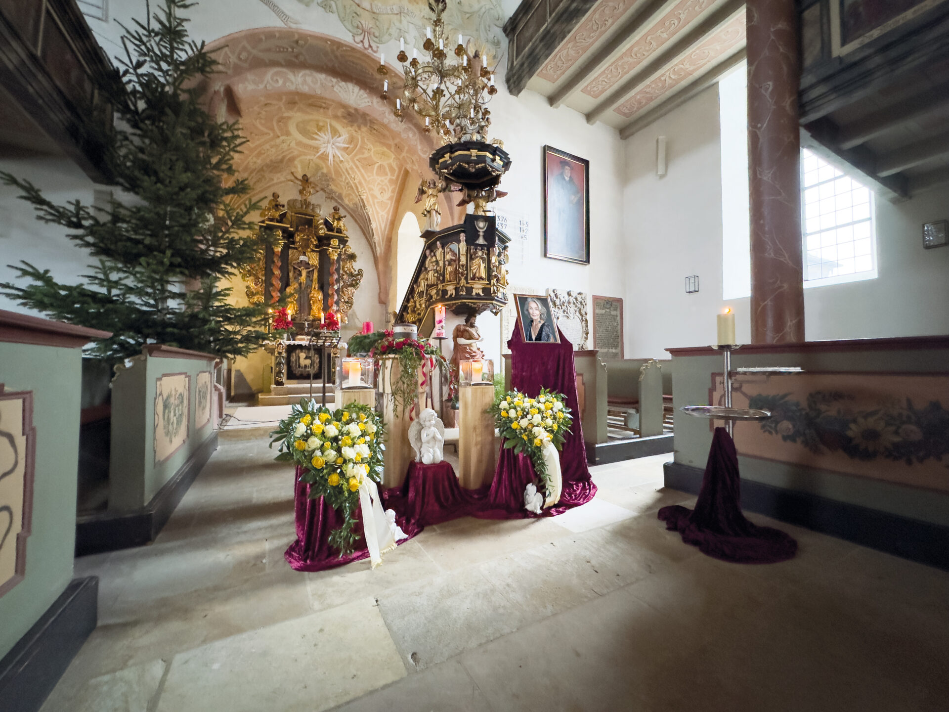 Ein geschmücktes Kircheninnere mit Blumenarrangements, Kerzen, einem Weihnachtsbaum, religiösen Statuen und einer reich verzierten Kanzel unter einer bemalten Decke, die eine festliche und ehrfürchtige Atmosphäre schafft.