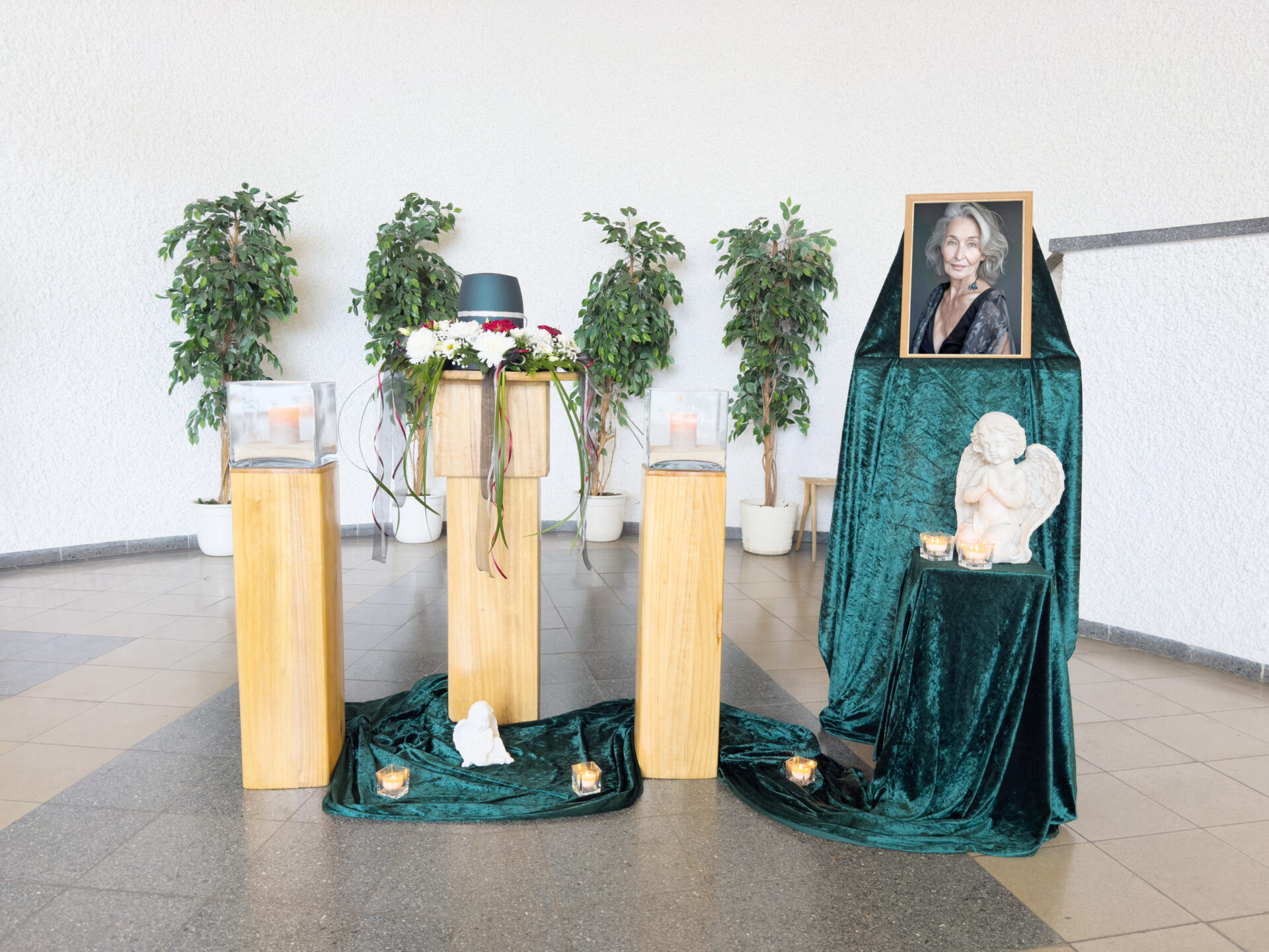 Ein Gedenkdisplay mit drei Holzsockeln, die Glaskästen, ein dunkelgrünes Tuch, Kerzen, eine weiße Engelsstatue und ein gerahmtes Foto einer lächelnden Frau enthalten, mit Topfpflanzen im Hintergrund.