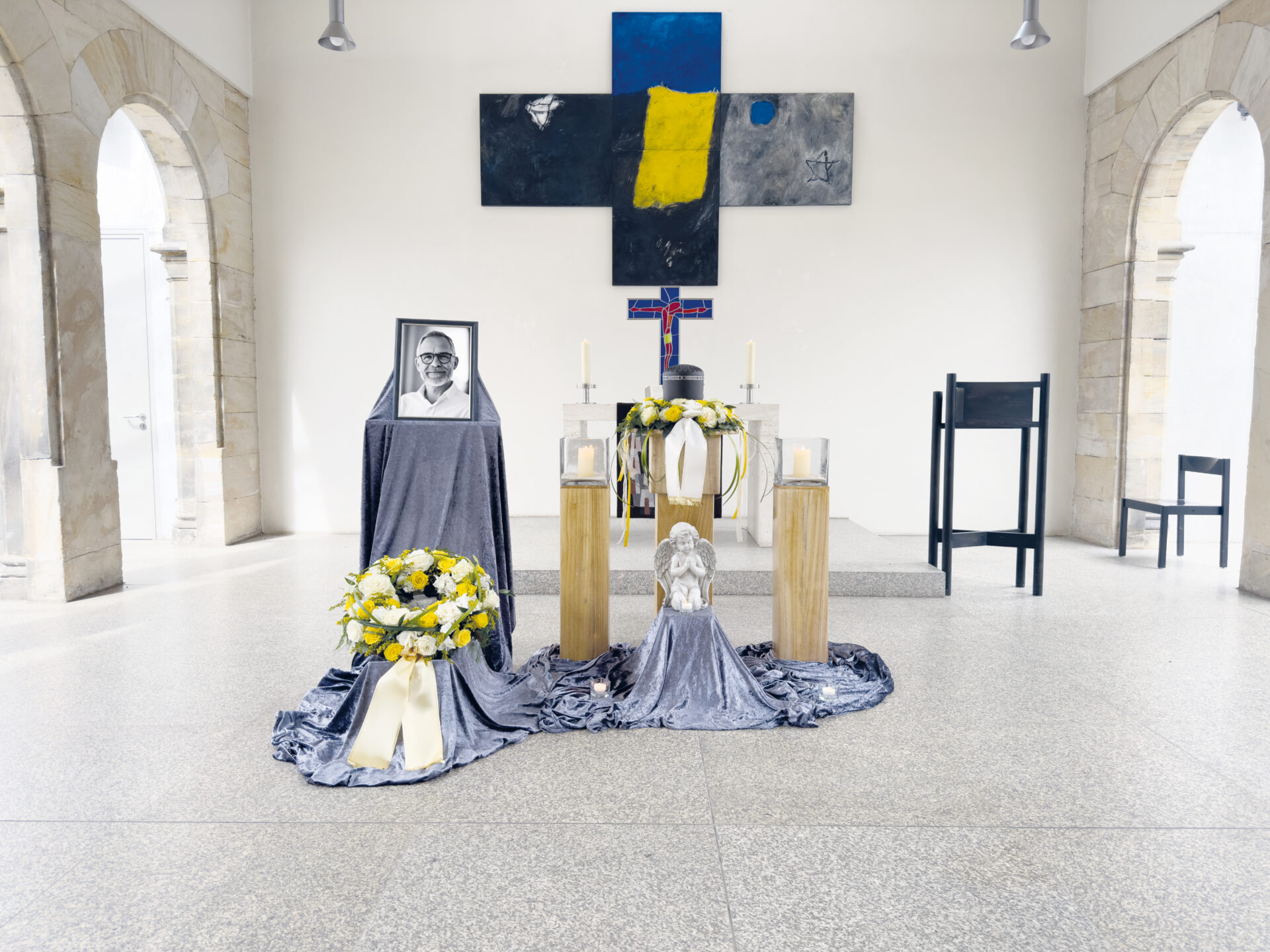 Eine Gedenkstätte mit einem gerahmten Schwarz-Weiß-Porträt, Blumenarrangements, Kerzen und einer Engelsfigur, die in einem hellen Raum mit gewölbten Steintüren und einem großen blau-gelben Kreuz an der Wand aufgestellt ist.