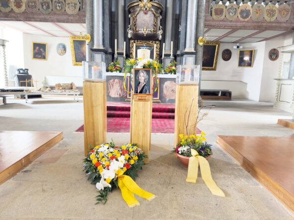 In einer Kirche ist das Porträt einer älteren Frau auf einem Holzständer zu sehen, umgeben von Blumenkränzen mit gelben Bändern und zwei kleineren Holzsockeln mit Glasbehältern.