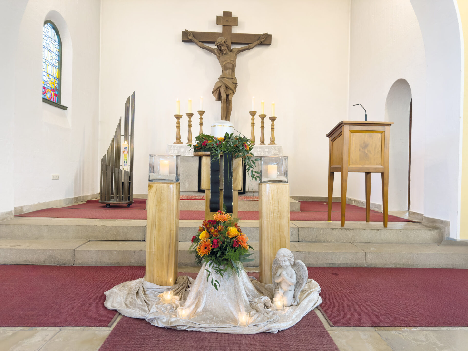 Ein Altar in einer Kirche mit einem Kruzifix an der Wand, umgeben von Kerzen, Blumen, zwei hohen Holzsockeln und einer kleinen Engelsfigur. Der Boden ist mit rotem Teppich ausgelegt, und rechts steht eine Kanzel.