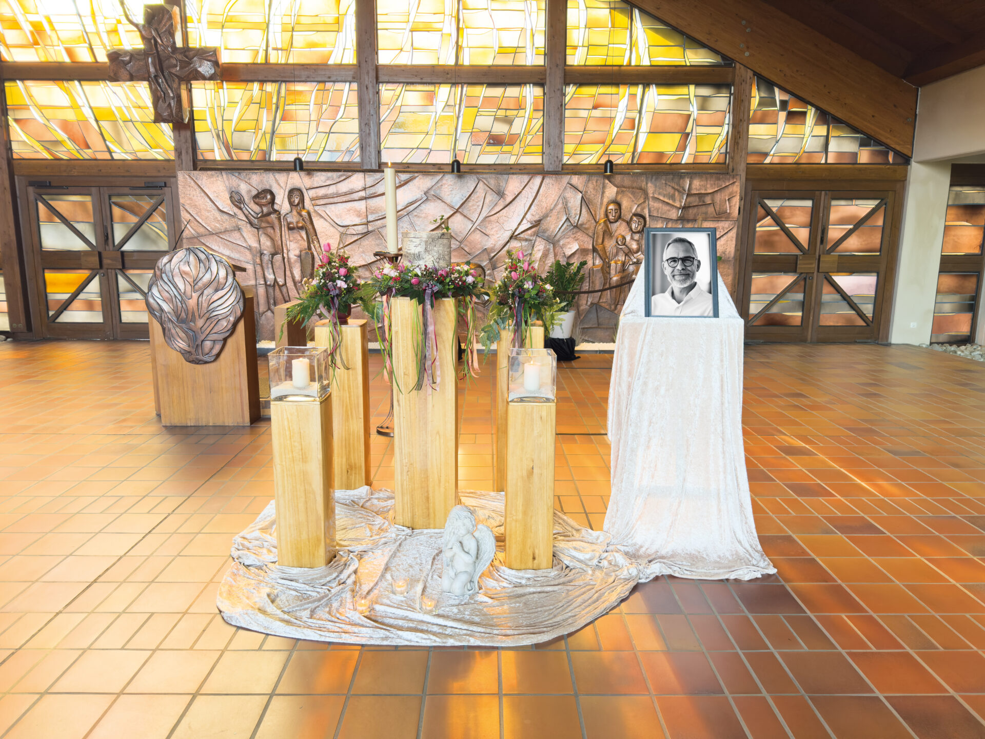 Ein Gedenkbild in einer Kirche zeigt Kerzen, Blumen und das Foto eines lächelnden Mannes auf weißem Stoff. Dahinter befinden sich Buntglasfenster und ein Bronzerelief mit menschlichen Figuren. Der Boden ist gefliest.