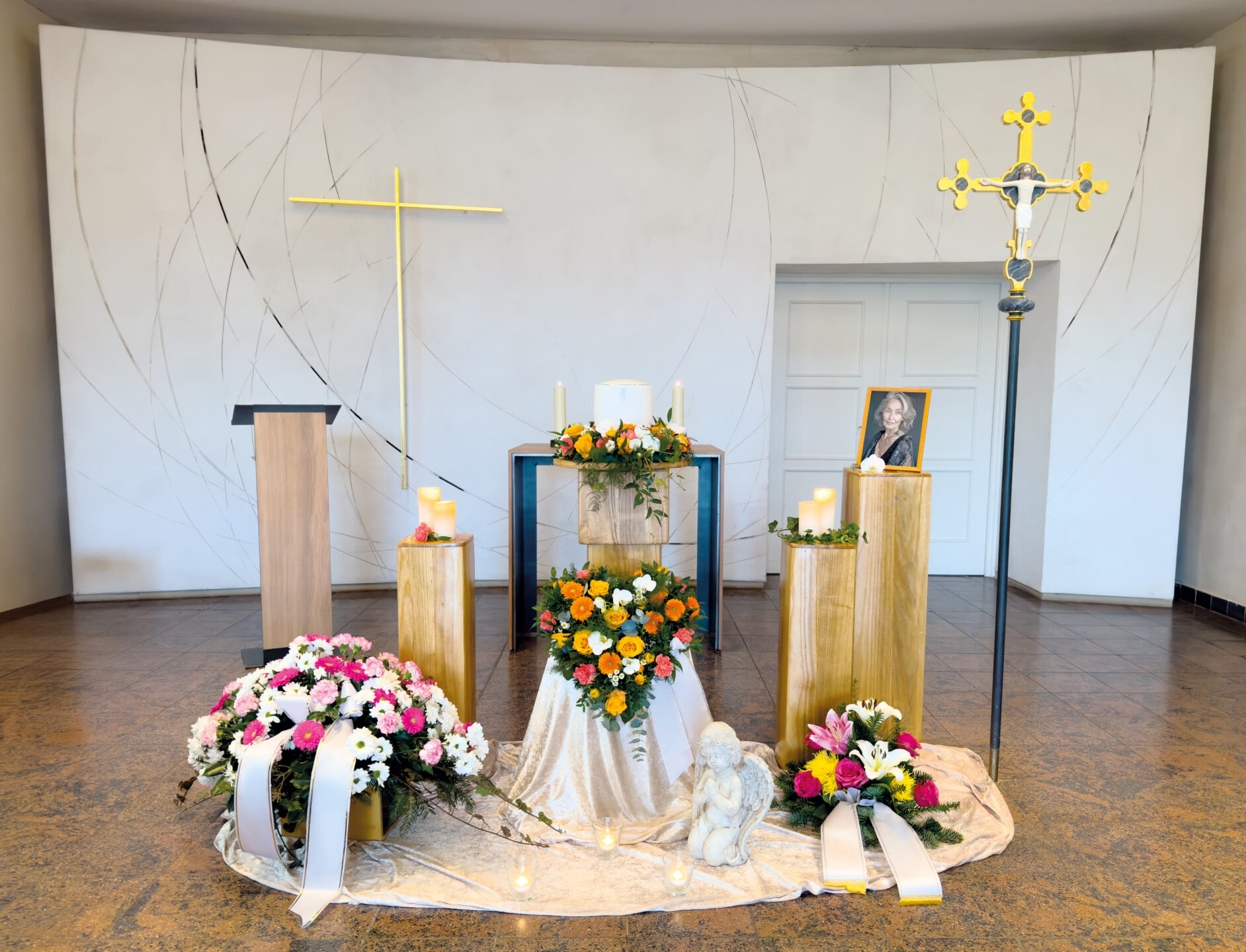 Eine Trauerfeier in einer Kapelle mit Blumenkränzen, Kerzen, einer Engelsfigur, einem goldenen Kreuz und einem gerahmten Foto einer Frau auf einem Sockel, mit einem großen Wandkreuz und einem Podium im Hintergrund.