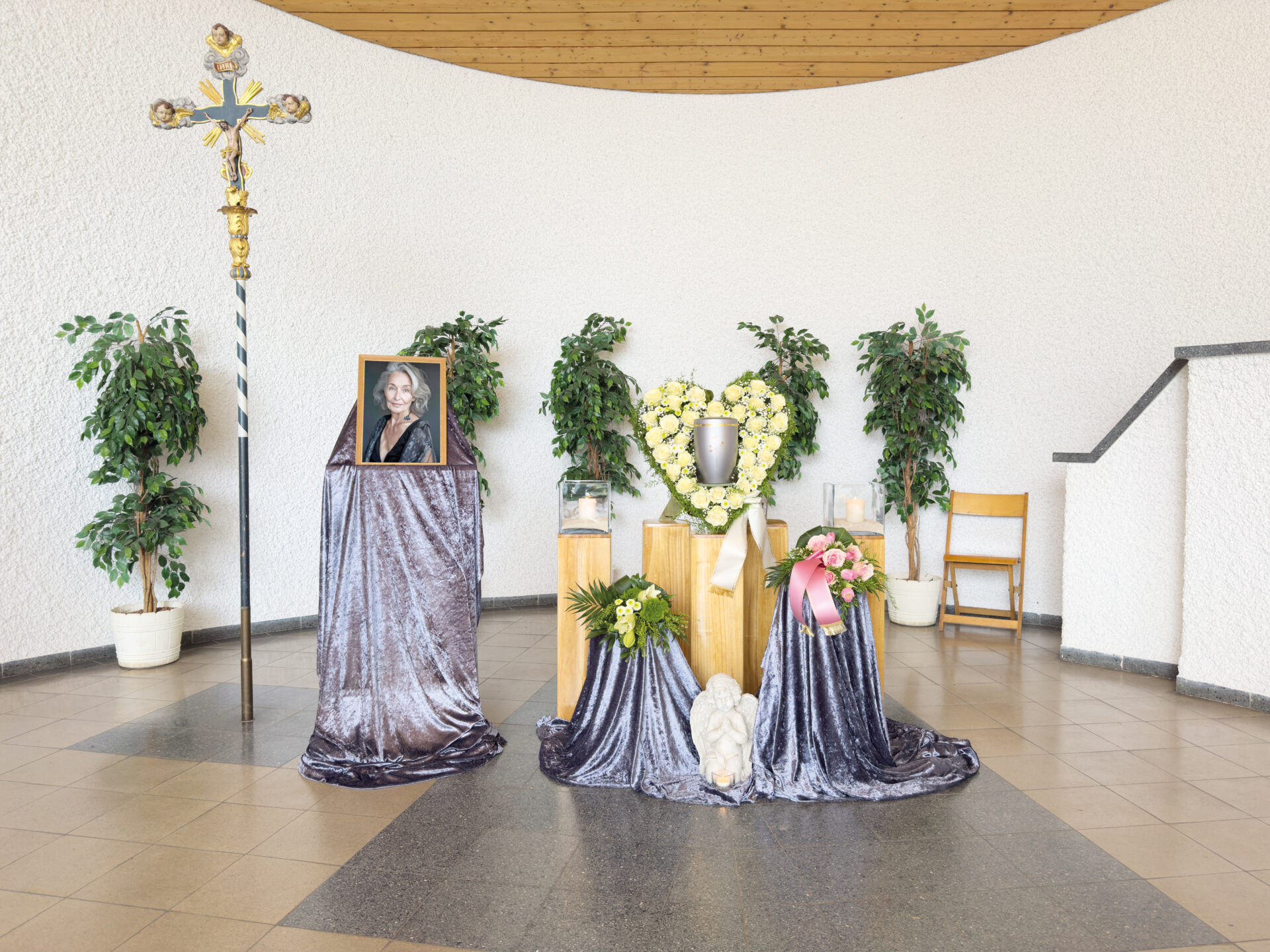 In einem hellen Raum ist eine Gedenkstätte mit einem gerahmten Porträt, einem Blumenherzkranz, Blumenarrangements, einem Kreuz und einem über Tische drapierten silbernen Stoff eingerichtet. Ein einzelner Holzstuhl steht an der geschwungenen weißen Wand.