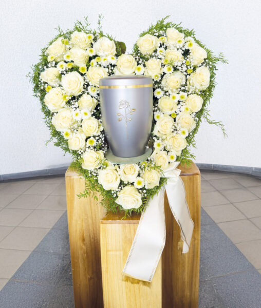 Eine silberne Urne mit Rosenmotiv steht auf einem Holzsockel, umgeben von einem herzförmigen Kranz aus weißen Rosen, Gänseblümchen, Grünzeug und einem weißen Band - perfekt für eine elegante Urnentrauerfeier.