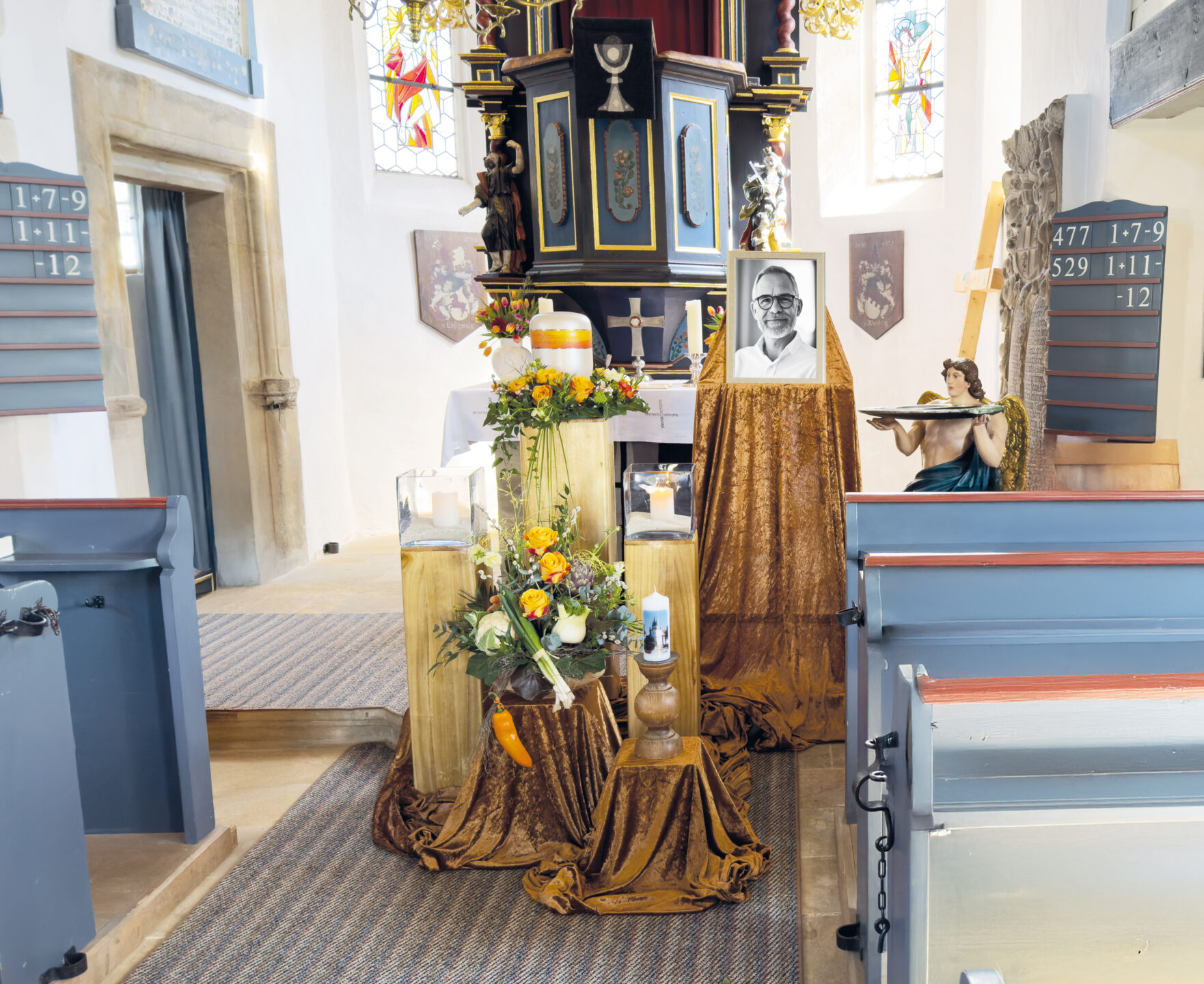 Ein mit Blumen, Kerzen und dem Porträt eines Mannes mit Brille geschmückter Kirchenaltar. Im Hintergrund sind Buntglasfenster und Gesangstafeln zu sehen, rechts stehen blaue Kirchenbänke und die Statue eines Engels, der ein Tablett hält.