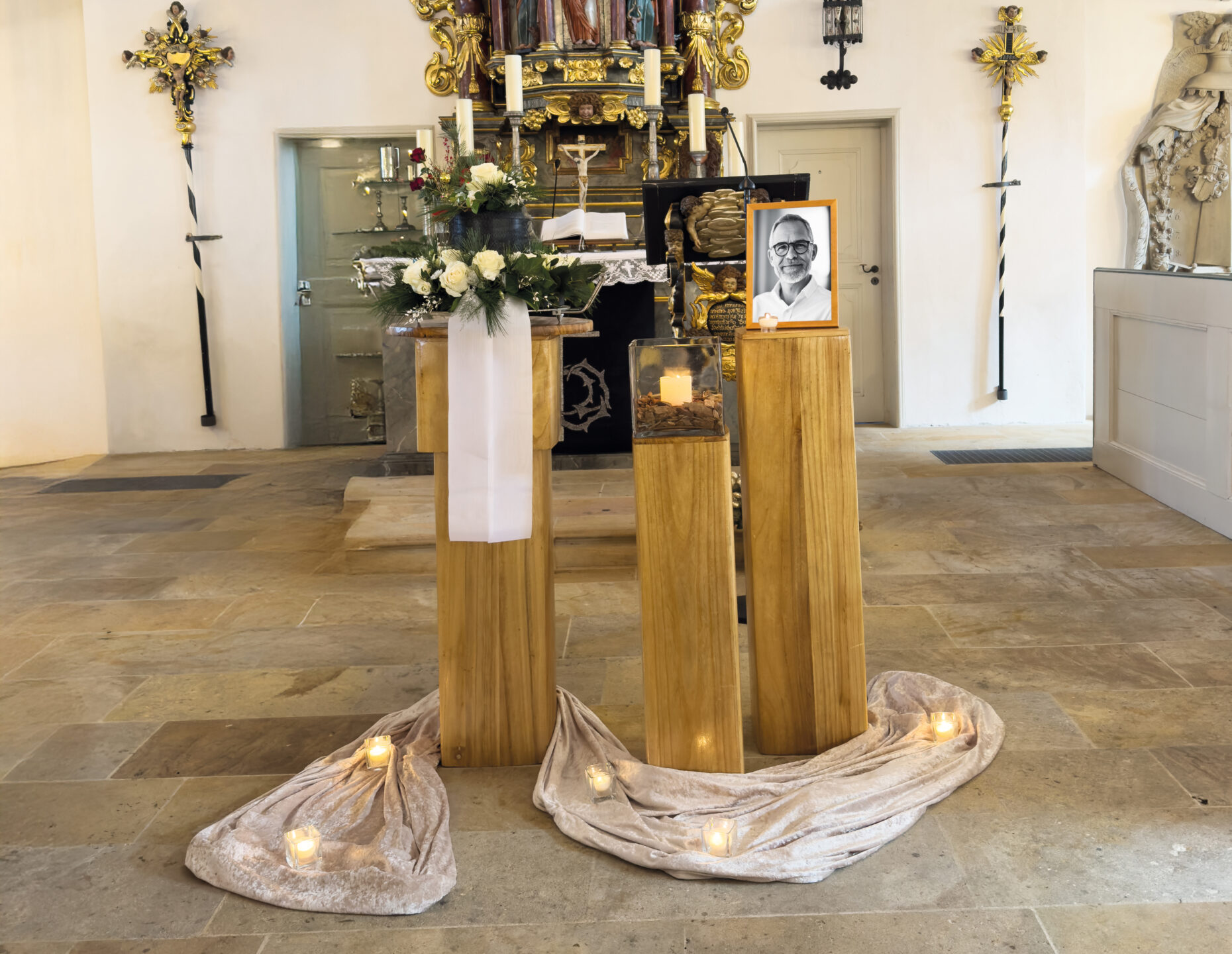 Eine in einer Kirche aufgestellte Gedenkstätte zeigt drei Holzsockel mit Blumen, Kerzen und einem gerahmten Schwarz-Weiß-Foto eines Mannes. Drapierter Stoff und Teelichter schmücken den Sockel. Im Hintergrund ist ein verschnörkelter Altar zu sehen.