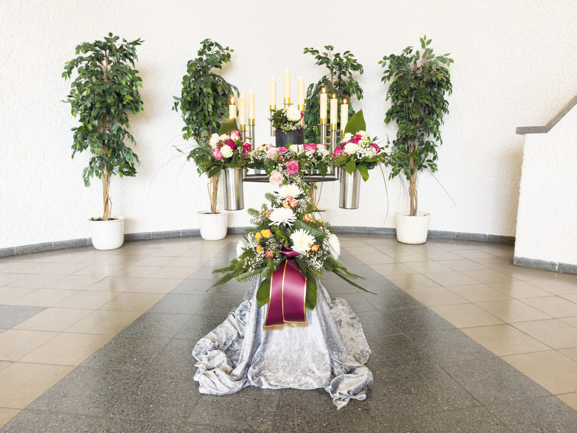 Ein Beerdigungsgesteck mit Blumensträußen, Kerzen und einem violetten Band auf einem gedeckten Tisch, flankiert von zwei grünen Topfpflanzen vor einem weißen Wandhintergrund.