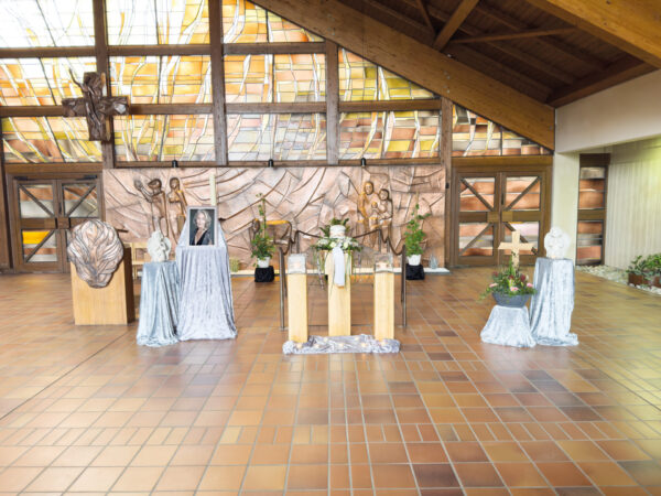 Ein Kirchenraum mit einer Gedenkausstellung: ein Foto einer Person, Kerzen, Blumen und Statuen auf Ständern mit silbernen Tüchern. Dahinter befindet sich eine Holzwand mit Skulpturen und einem Buntglasfenster.