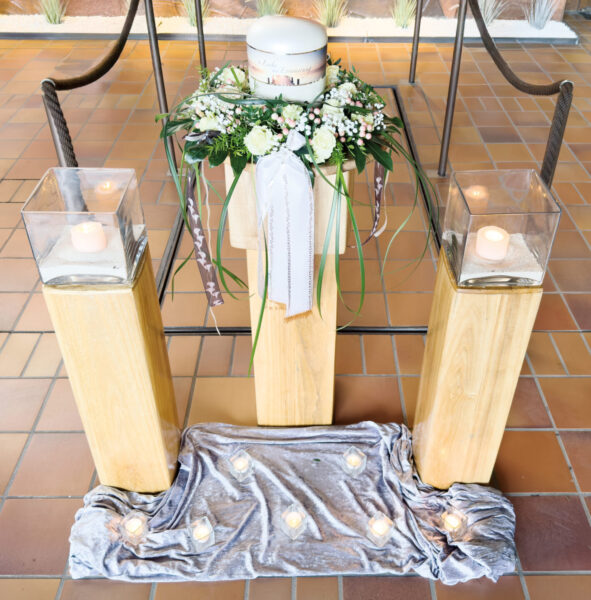 Eine weiße Urne mit Blumenschmuck und einer Schleife steht auf einem Holzständer und bildet einen eleganten Rahmen für die Urnentrauerfeier. Die brennenden Kerzen auf den Sockeln und der graue Stoff, der auf dem Boden drapiert ist, verleihen dem Arrangement Wärme.