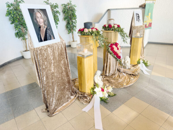Ein Gedenkdisplay für eine Urnentrauerfeier besteht aus gerahmten Fotos, einer Urne, Blumenkränzen, Kerzen und Gestecken mit weißen Bändern auf goldenen Ständern, die mit Samttüchern drapiert sind, in einem hellen Innenraum mit Topfpflanzen im Hintergrund.