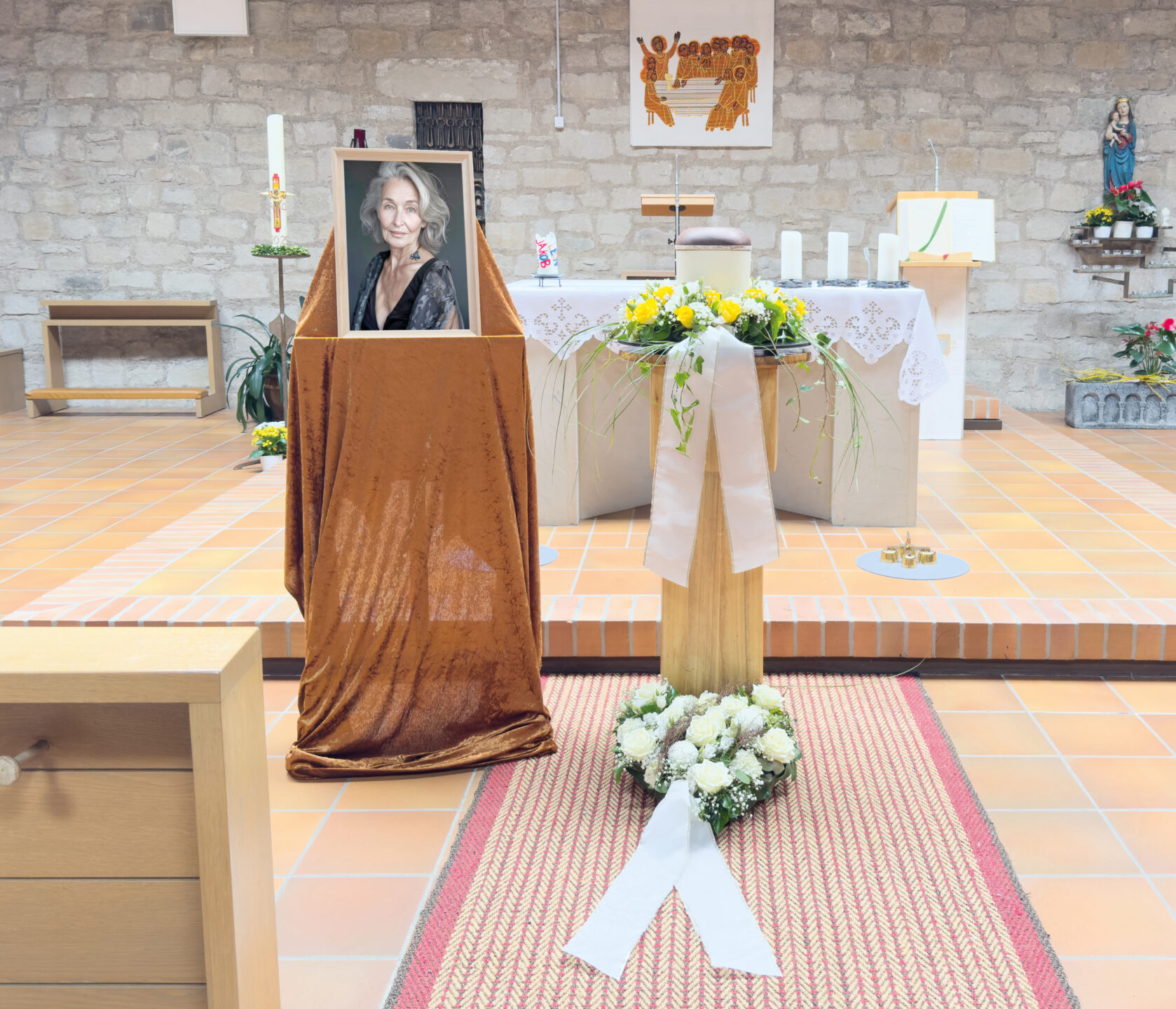 Ein Gedenkgottesdienst in einer Kirche mit dem gerahmten Foto einer älteren Frau auf einem mit braunem Stoff drapierten Ständer, einem Blumengesteck, einem weißen Band und einer Urne, mit einem Altar und religiösem Dekor im Hintergrund.