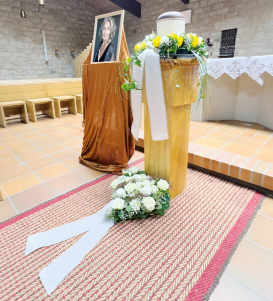 Ein Gedenkaufbau in einer Kirche für eine Urnentrauerfeier mit dem Porträt einer Frau, einer blumengeschmückten Urne auf einem Holzständer und einem Blumengesteck mit weißen Bändern auf einem rot gemusterten Teppich. Im Hintergrund befinden sich Holzbänke und ein Altar.