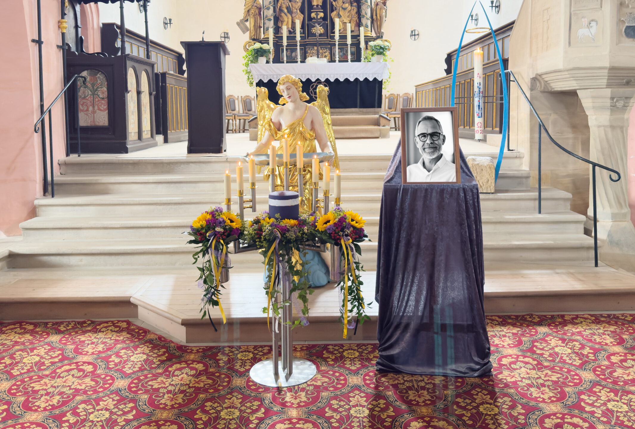 Eine Gedenkstätte in einer Kirche zeigt ein gerahmtes Schwarz-Weiß-Porträt auf einem drapierten Ständer, ein hohes Kerzenarrangement mit Blumen und eine goldene Engelsskulptur, die alle vor dem Altar auf einem rot gemusterten Teppich stehen.