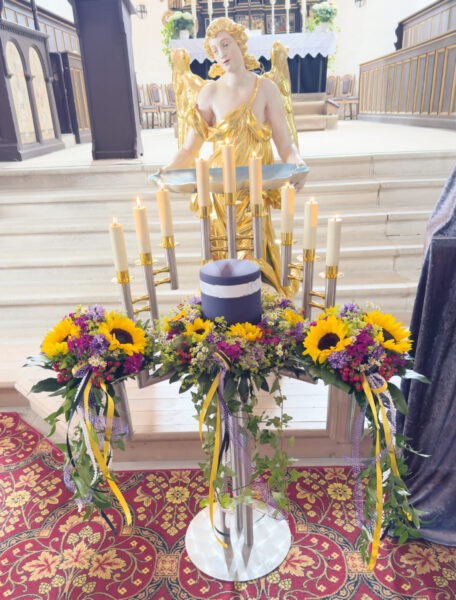 Eine Gedenkanzeige für eine Urnentrauerfeier zeigt eine blaue Urne, umgeben von gelben Sonnenblumen und violetten Blumen, die vor einer goldenen Engelsstatue und brennenden Kerzen in einer Kirche mit kunstvollem Dekor steht.