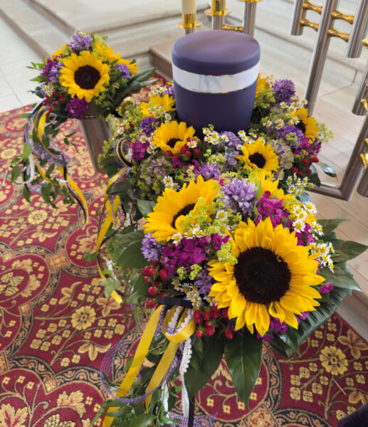 Eine violette Urne, das Herzstück der Urnentrauerfeier, ist von farbenfrohen Trauergestecken - Sonnenblumen und gelbe, violette und weiße Blüten - umgeben, die auf einem dekorativen, rot und gold gemusterten Teppich ruhen.