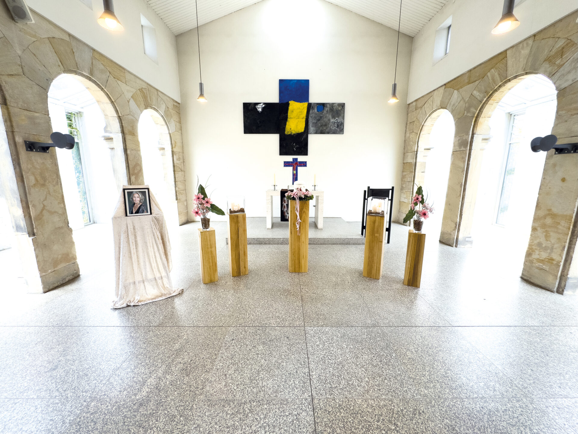 Das Innere einer Kapelle mit hohen Bogenfenstern, einem modernen Altar, Blumenarrangements, einem gerahmten Foto auf einem drapierten Ständer und fünf davor angeordneten Holzpodesten. An der Rückwand hängen ein Kreuz und ein abstraktes Kunstwerk.