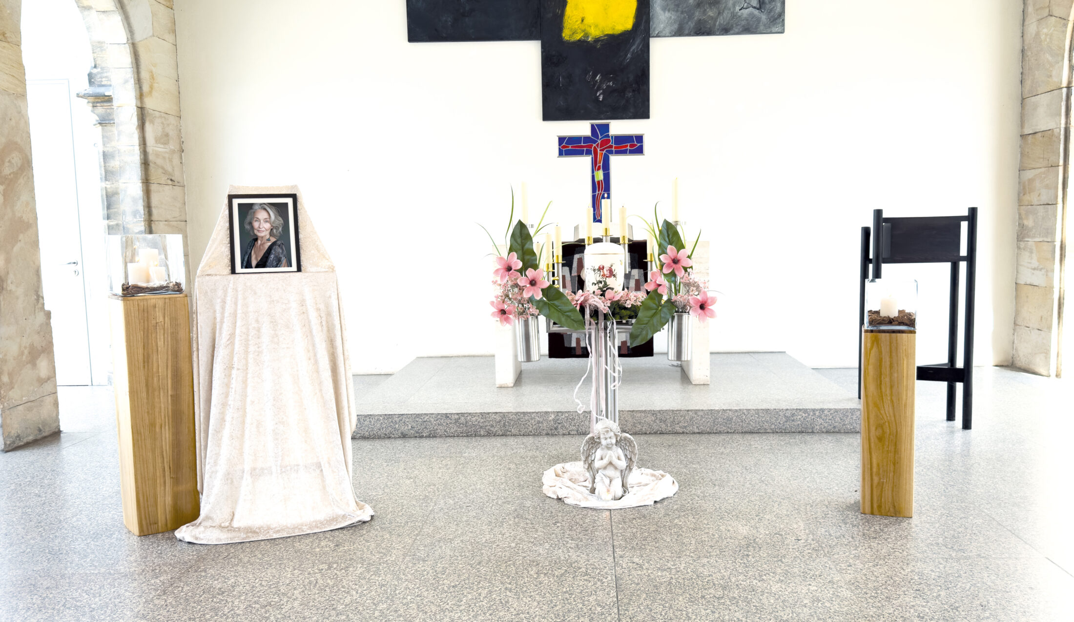 Eine Gedenkstätte in einer hellen Kapelle bietet ein gerahmtes Foto auf einem drapierten Ständer, Blumenarrangements, Kerzen und ein Kreuz auf dem Altar, über dem ein modernes Kunstwerk hängt.