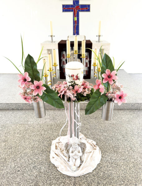 Ein dekorativer Kirchenaltar für eine Urnentrauerfeier zeigt eine große weiße Kerze mit floralen Motiven, Kunstblumen, rosa Blumen und Grünzeug in Metallvasen. Sechs goldene Kerzenhalter und ein Kreuz mit blauen und roten Details stehen hinter einer kleinen Engelsstatue.