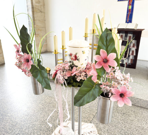 Eine weiße Urne mit floralen Motiven steht auf einem Ständer, umgeben von rosa Kunstblumen und Grünzeug. Im Hintergrund einer hellen Kapelle sind Kerzen und ein Altar zu sehen.