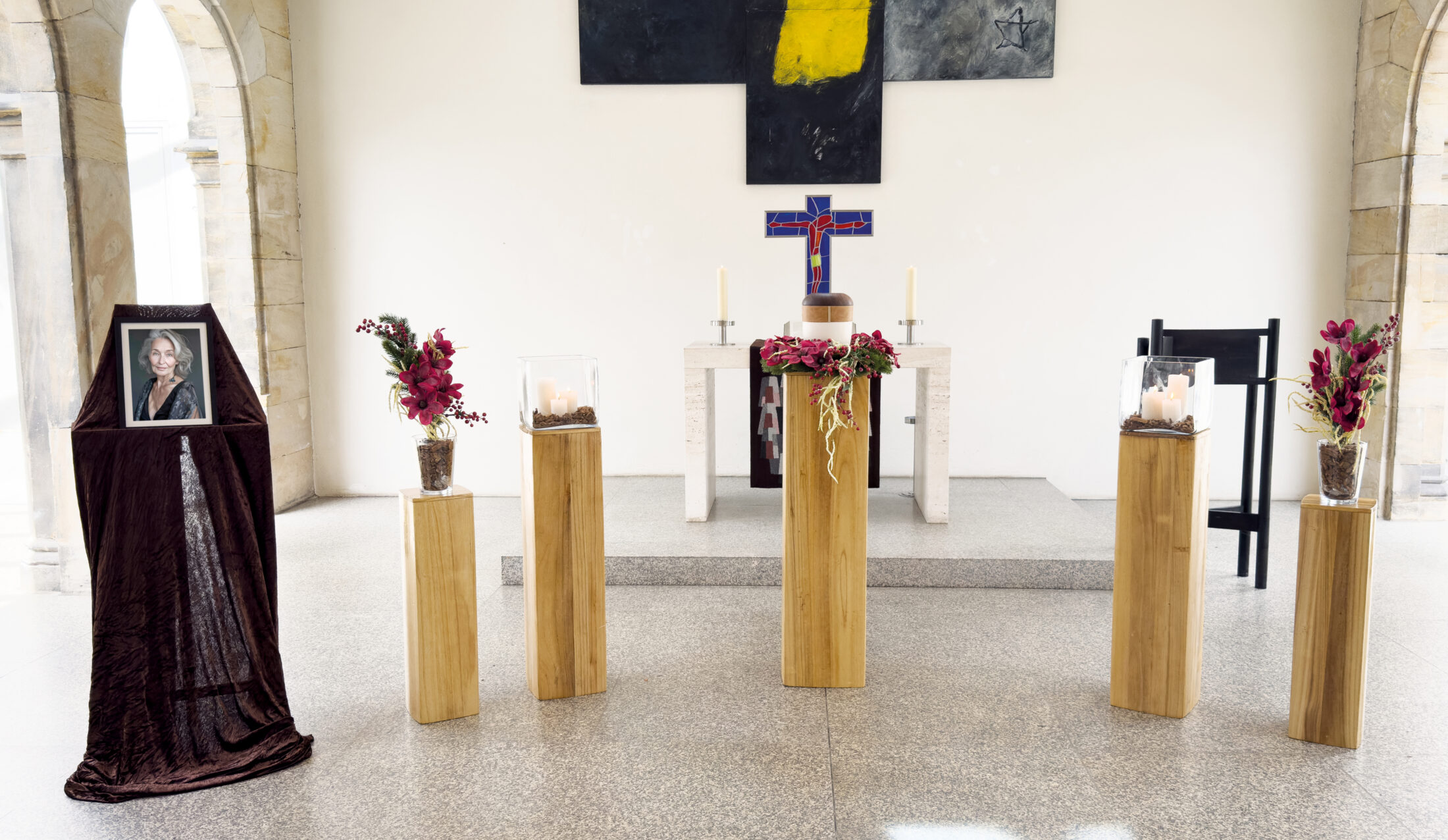 Eine Gedenkstätte mit fünf hölzernen Ständern mit Blumen und Kerzen, einem gerahmten Foto einer Frau auf einem drapierten Ständer und einem Altar im Hintergrund mit Kerzen, einem Kreuz und abstrakter Kunst an der Wand.