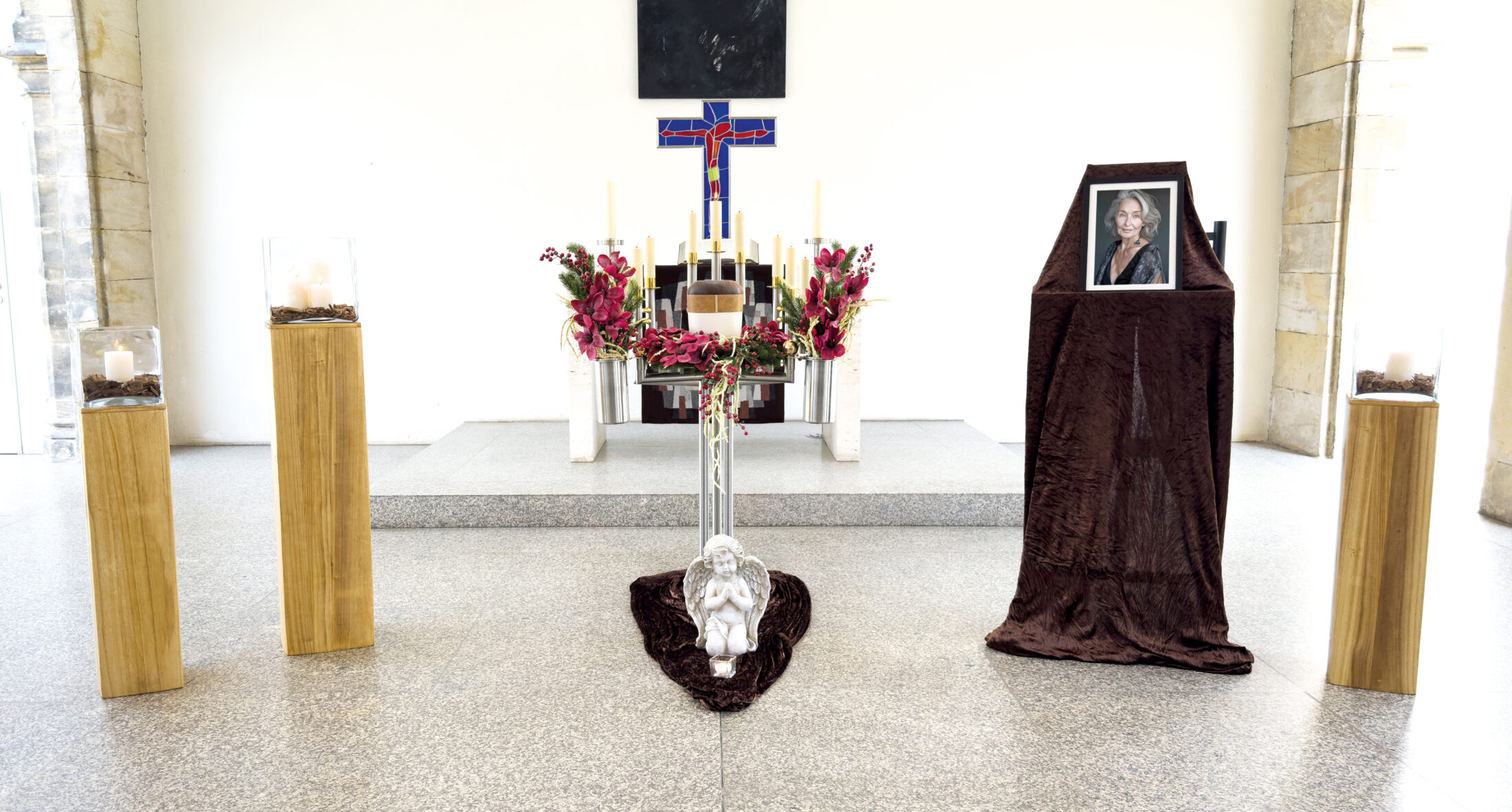 Ein Begräbnisaltar mit Kerzen, Blumen, einem Kreuz und einer Engelsstatue. Auf der rechten Seite ruht ein gerahmtes Foto einer Frau auf einem mit braunem Stoff bezogenen Ständer. Die Umgebung ist hell, das Licht strömt herein.