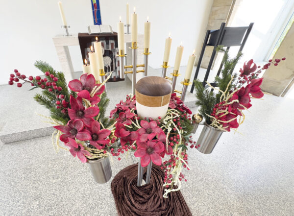 Eine weiße Urne ist umgeben von roten Blumen, Kunstblumen, grünem Laub und Kerzen auf Metallständern in einem hellen Kirchenraum während einer Urnentrauerfeier, mit einem Altar, einer Bibel und weiteren Kerzen im Hintergrund.