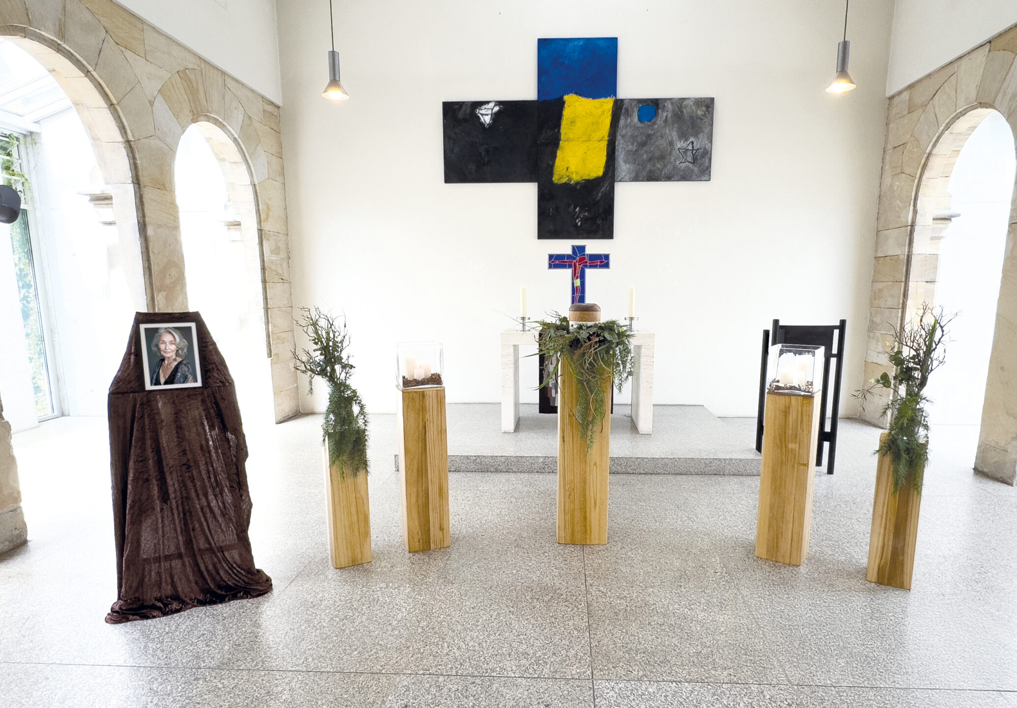 Eine Kapelle mit Steinbögen, fünf Holzsockeln mit Grünzeug, einem drapierten Fotoporträt und einem schwarzen, kreuzförmigen Kunstwerk in Gelb und Blau über dem Altar. Auf dem Altartisch stehen Kerzen und ein kleines Kreuz.
