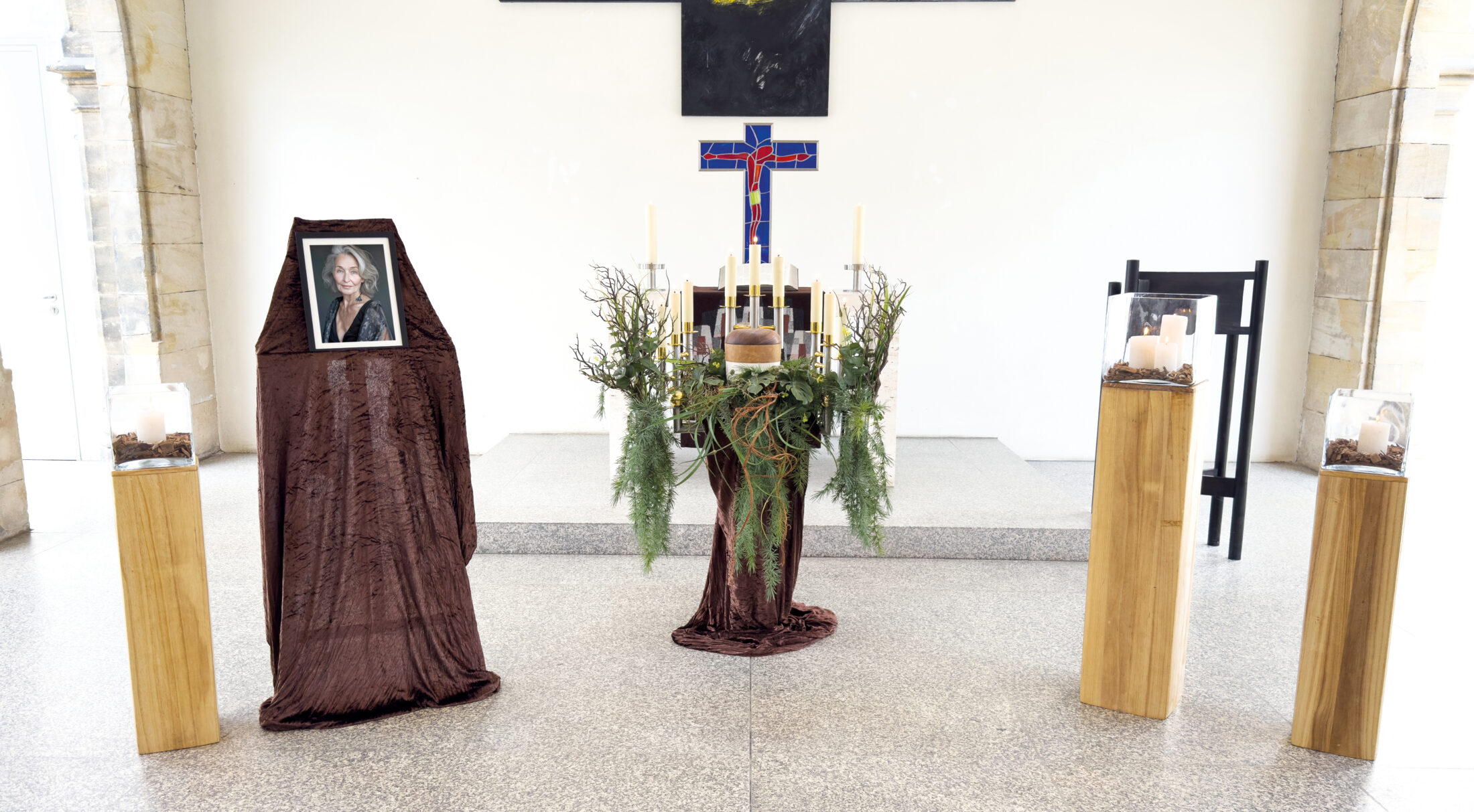 In einem hellen Raum steht ein mit braunem Stoff bespannter Ständer mit einem gerahmten Foto, einem Kerzengesteck mit Grünzeug und zwei Sockeln mit Glaskästen mit Kerzen. An der Wand dahinter hängt ein Kreuz.