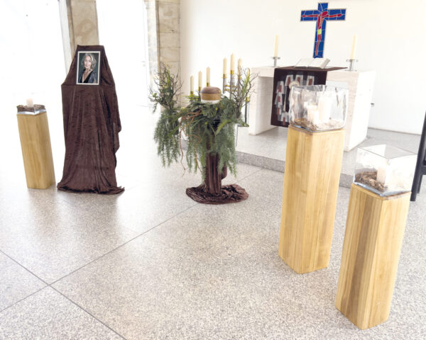 In einem hellen Raum findet eine Urnentrauerfeier statt, mit einem gerahmten, in braunes Tuch gehüllten Foto, Kunstblumen und einer Urne mit Blattwerk, Kerzen und drei Holzpodesten mit Gegenständen in Vitrinen neben einem Altar mit Kreuz.