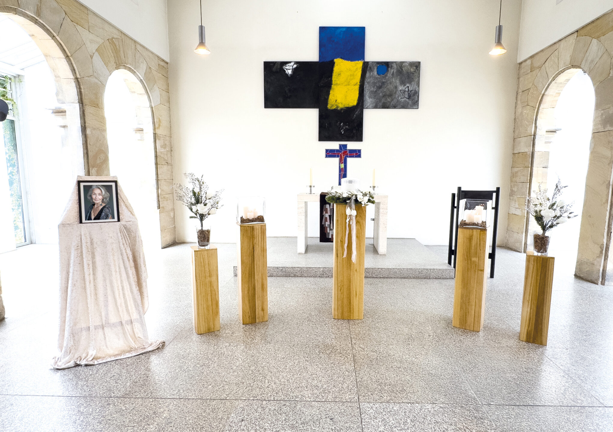 Ein Kapellenaltar mit vier hölzernen Sockeln, auf denen weiße Blumengestecke stehen, ein Foto einer Frau auf der linken Seite und ein schwarz-gelbes Kreuz an der Wand hinter dem Altar.