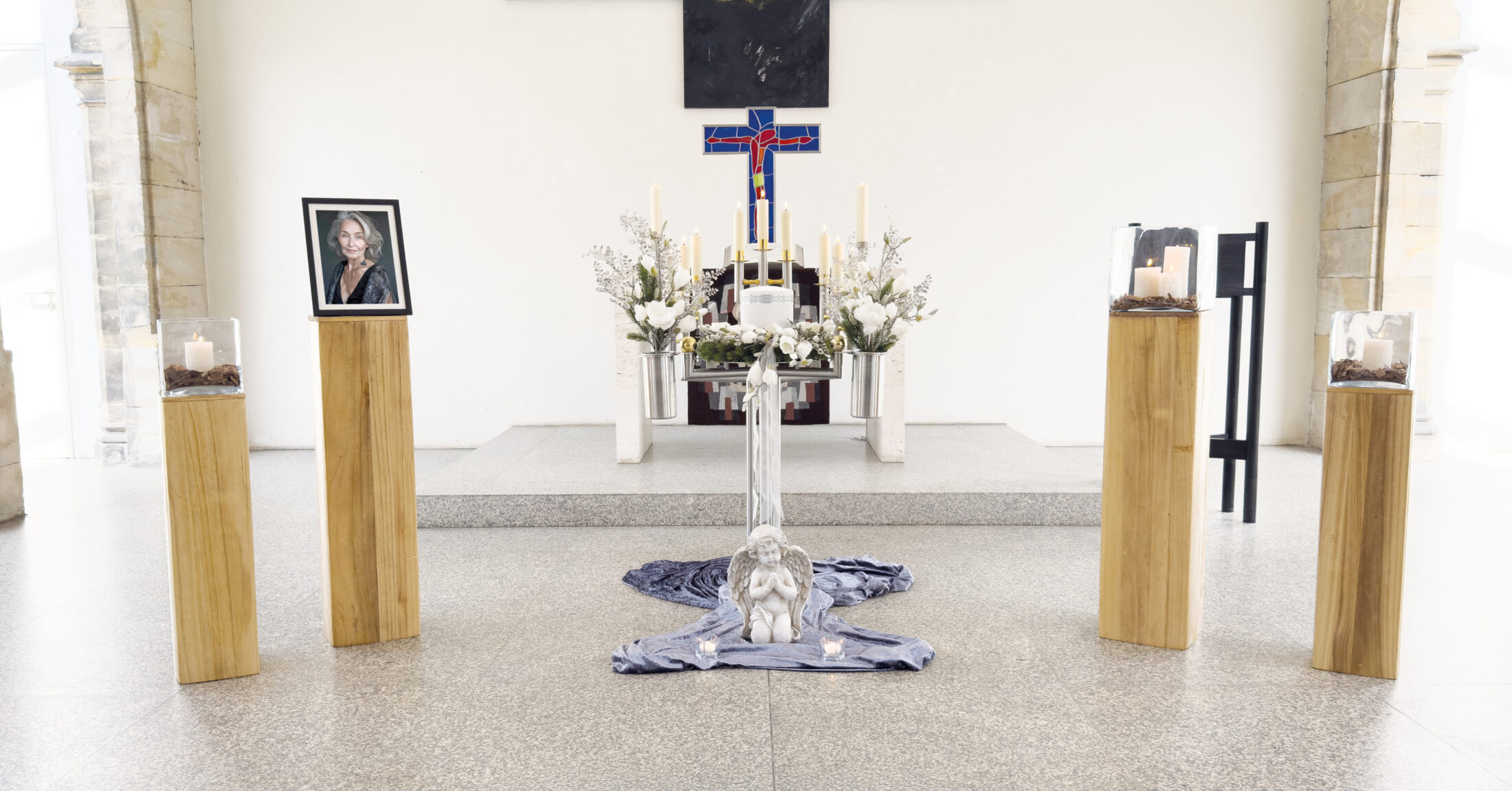 Eine Gedenkstätte in einer Kirche mit einem gerahmten Foto auf einem Holzsockel, Kerzen, weißen Blumen, einem Kreuz und einer kleinen Engelsstatue in der Mitte, die eine ruhige und respektvolle Atmosphäre schafft.