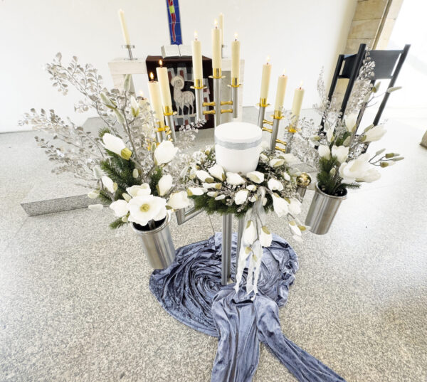 Eine weiße Urne, umgeben von Kunstblumen, silbernem Laub und neun brennenden Kerzen auf Ständern, alles arrangiert auf einem blauen Samttuch in einer hellen, modernen Urnentrauerfeier für den Innenbereich.