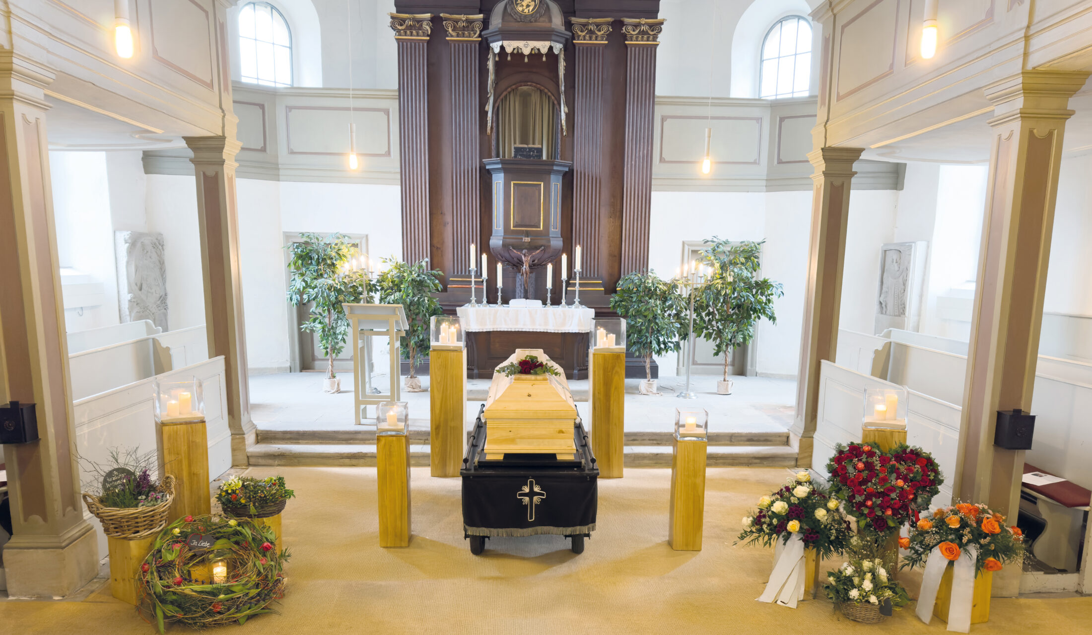 Ein hölzerner Sarg mit einem Blumengesteck steht während einer Trauerfeier vor einem Kirchenaltar, umgeben von Kerzen, Kränzen und Blumen. Die Kirche hat weiße Wände, Säulen und hohe Fenster.